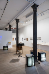 Color photograph of a gallery space. Along the middle of the space are placed various television monitors and musical instruments and audio devices. To the right on the wall are a series of photographs and prints, and two vitrines hang on the wall.