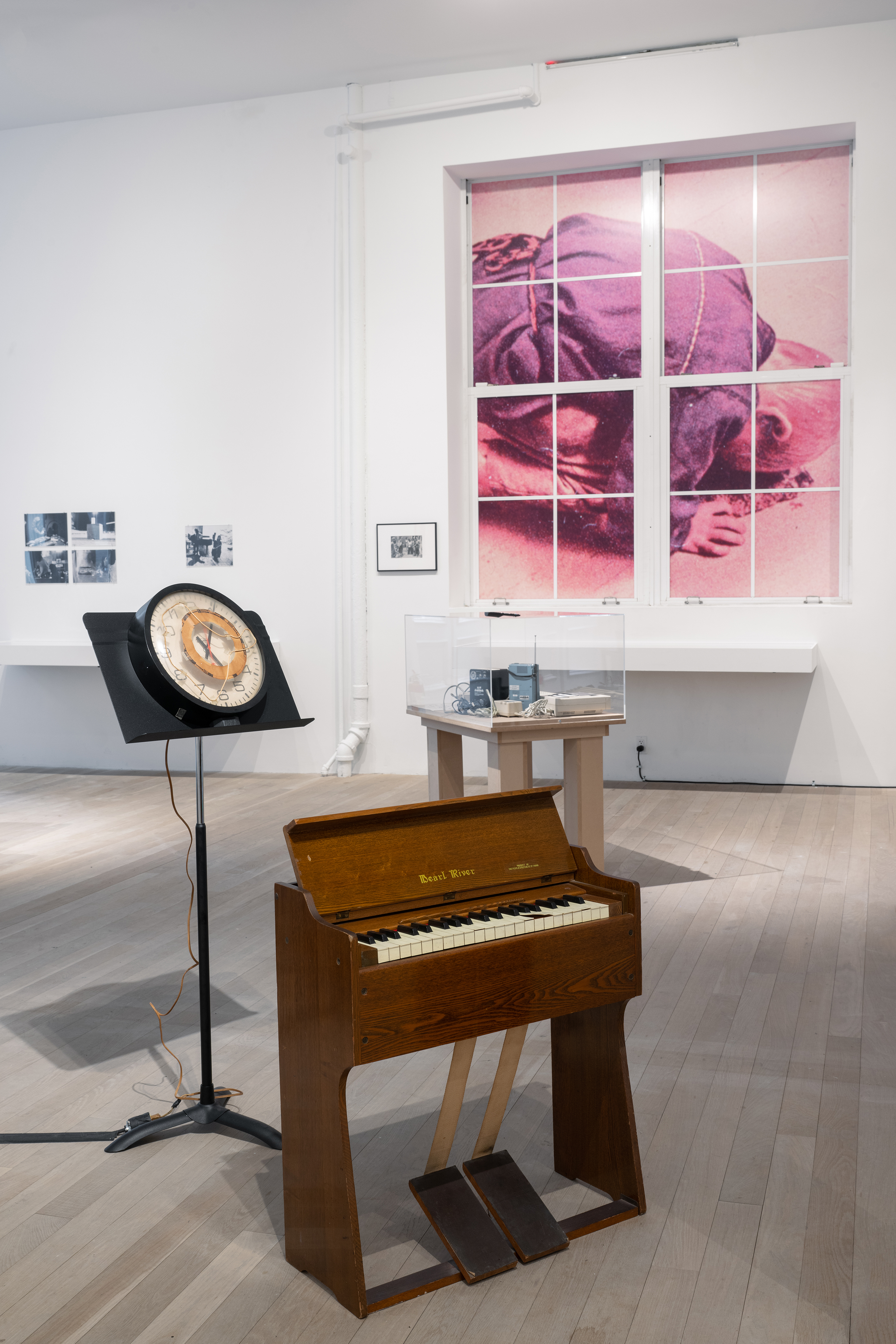 Color photograph of a gallery space. In the foreground is a small reed organ, with a music stand holding a clock device behind it. In the background are vitrines holding other audio devices and ephemera. On the window to the right is a blown-up image of a man kneeling with his head to the floor. Black-and-white photographs hang on the wall.