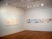 A corner of a gallery space with wooden floors. Two framed works on paper hang on the left wall. A light blue color has also been imprinted on the central portion of the wall. A long scroll of paper stretches horizontally across the wall to the right. The paper is covered with black and pastel-colored markings.