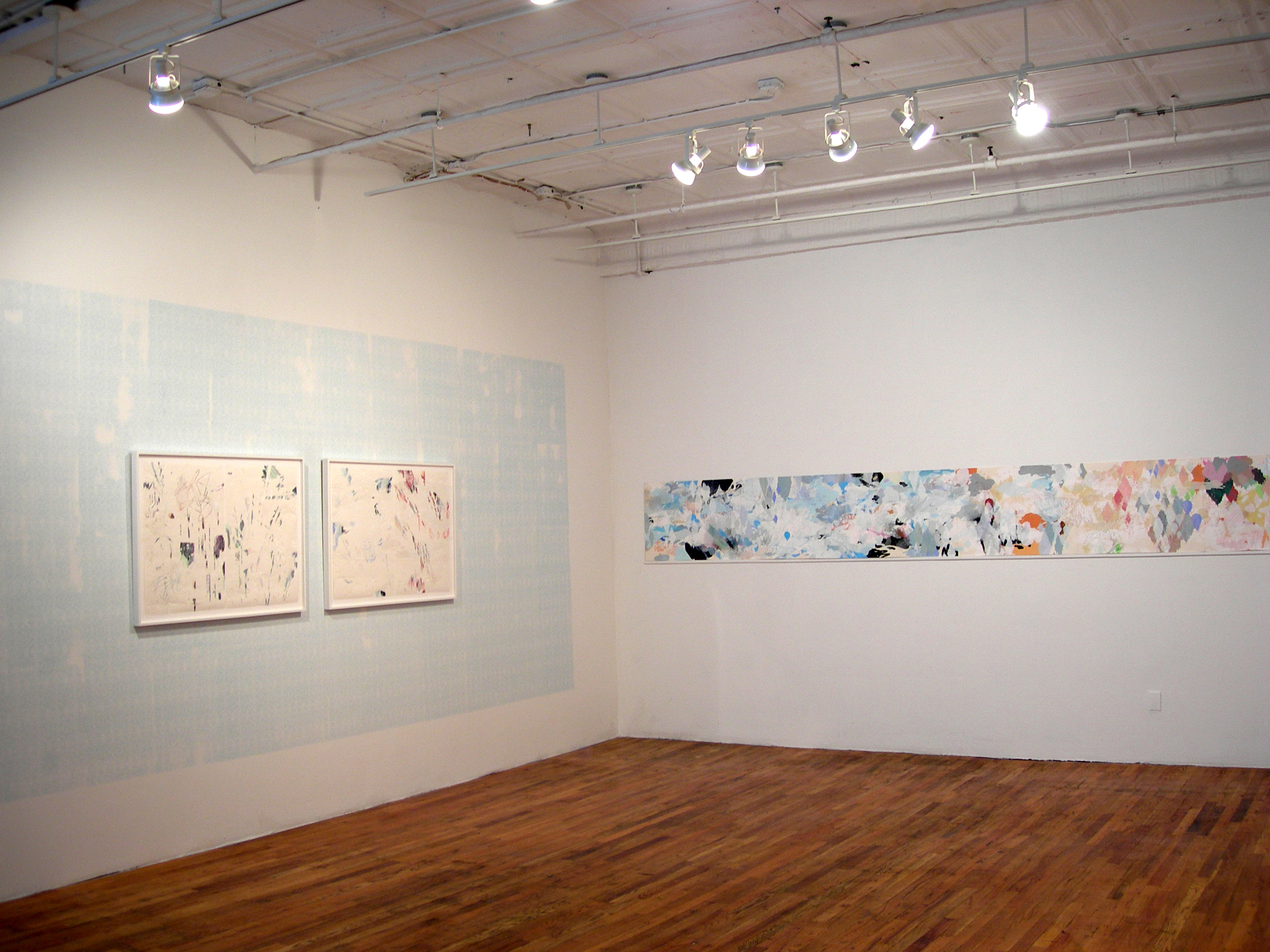 A corner of a gallery space with wooden floors. Two framed works on paper hang on the left wall. A light blue color has also been imprinted on the central portion of the wall. A long scroll of paper stretches horizontally across the wall to the right. The paper is covered with black and pastel-colored markings.