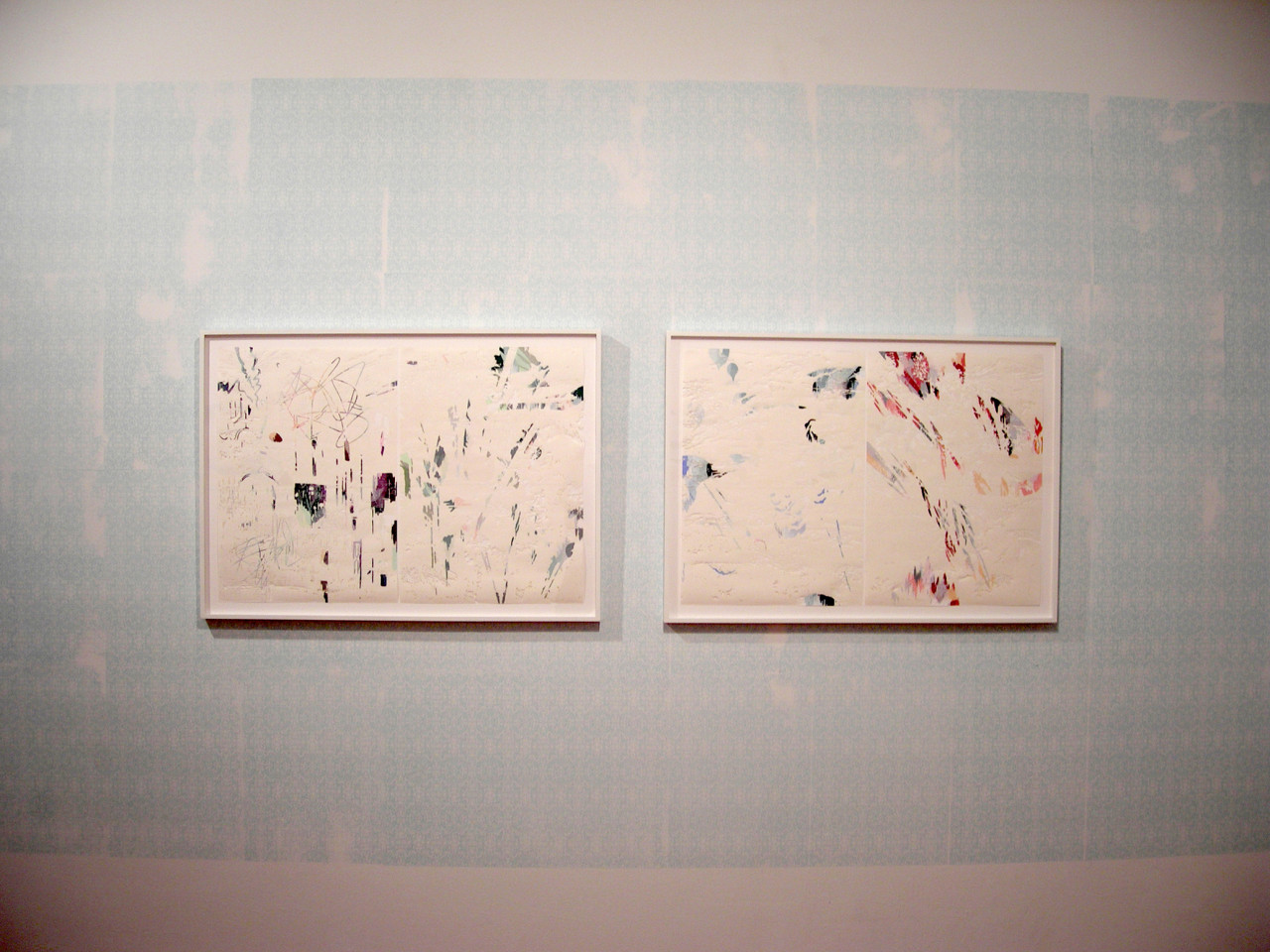 Two delicate, framed works on paper hang side by side against a white wall. A light blue color has been imprinted on the central portion of the wall. The artworks feature flecks of pastel color on an otherwise white surface.