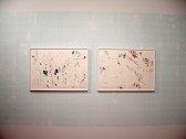 Two delicate, framed works on paper hang side by side against a white wall. A light blue color has been imprinted on the central portion of the wall. The artworks feature flecks of pastel color on an otherwise white surface.