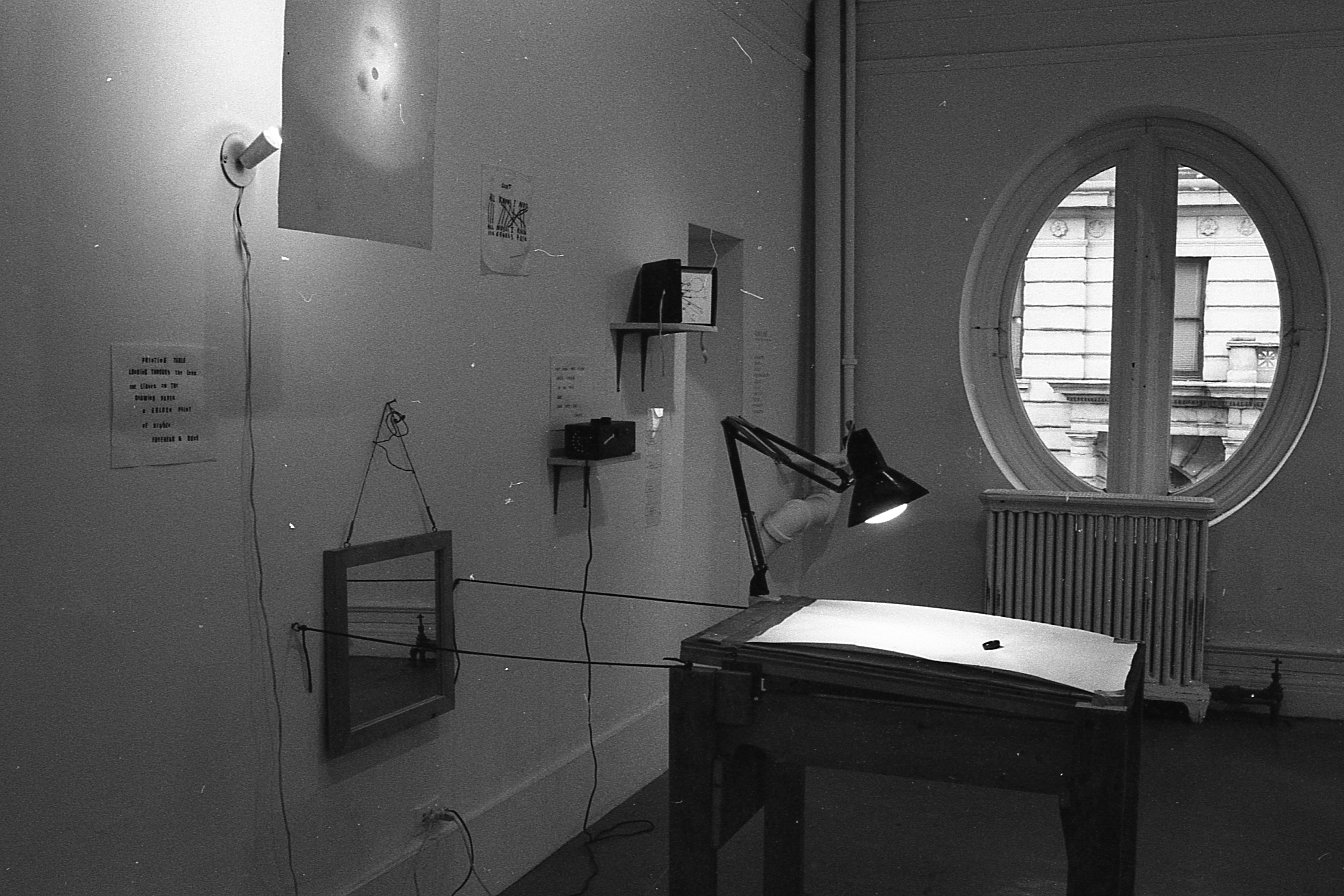 A number of objects and writings appear on the gallery wall and a desk with a light appears in front of it with windows visible in the background.