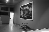 A gallery space with a chalk drawing on the top section of a wall and chairs underneath. Framed images on the adjacent wall are near a doorway into another space.
