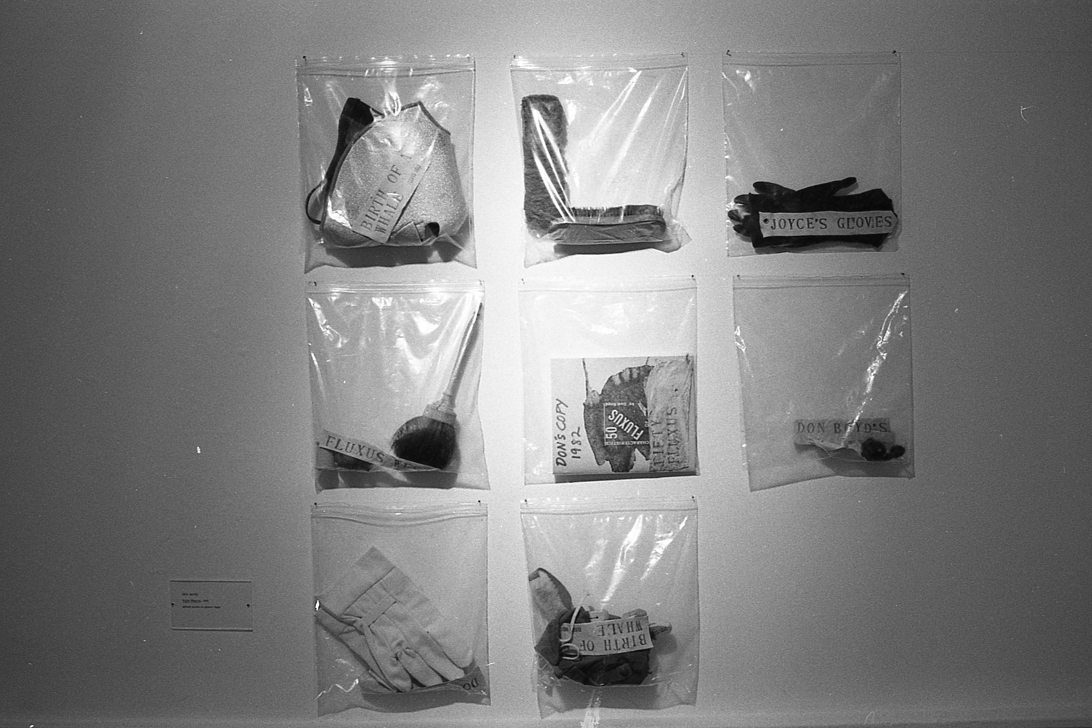 Plastic bags of objects appear on a white gallery wall.