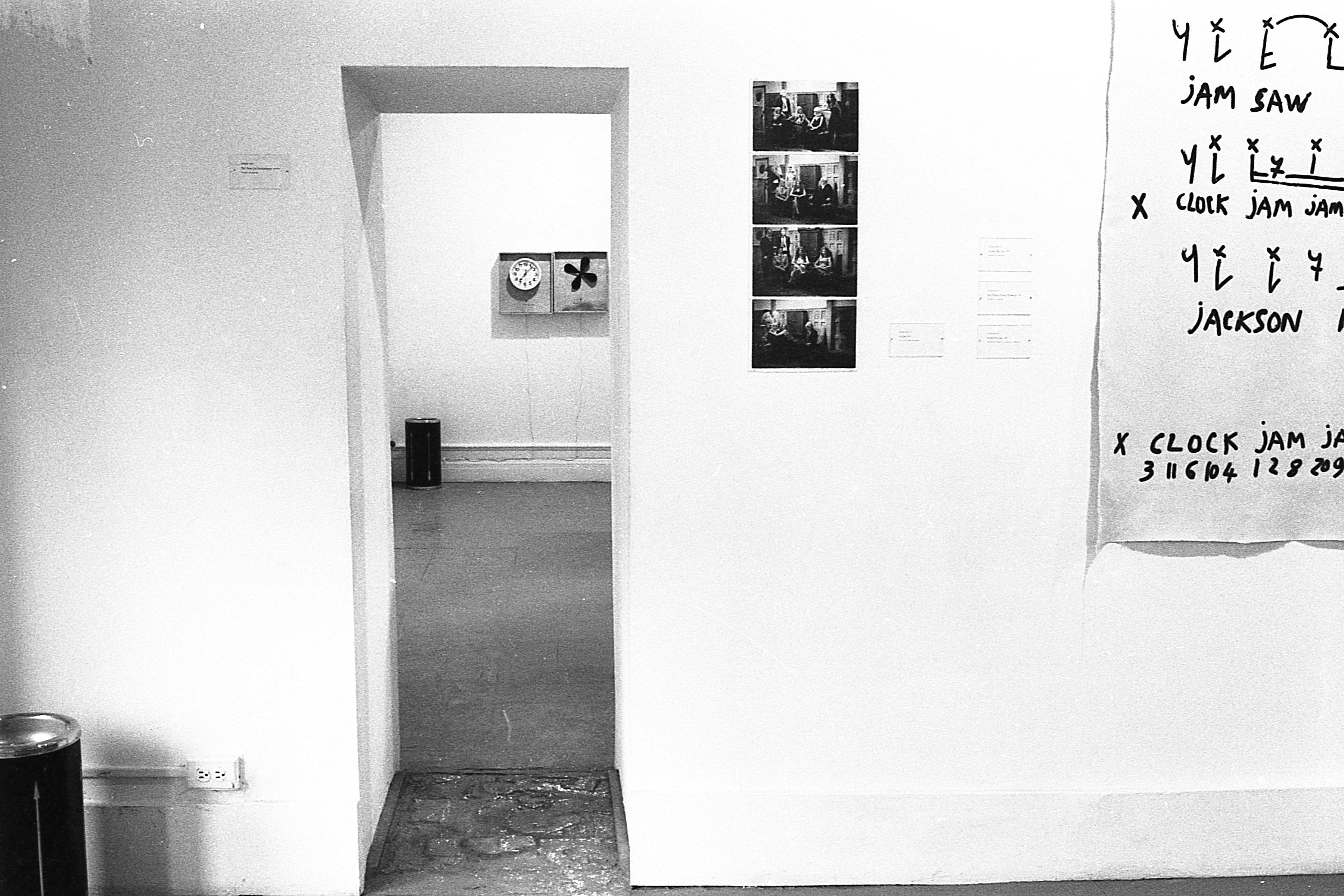 View through a doorway with photographs and a black text on a poster on the foreground wall and a clock along with a propeller of fan upholstered to the wall on the back wall through the doorway.