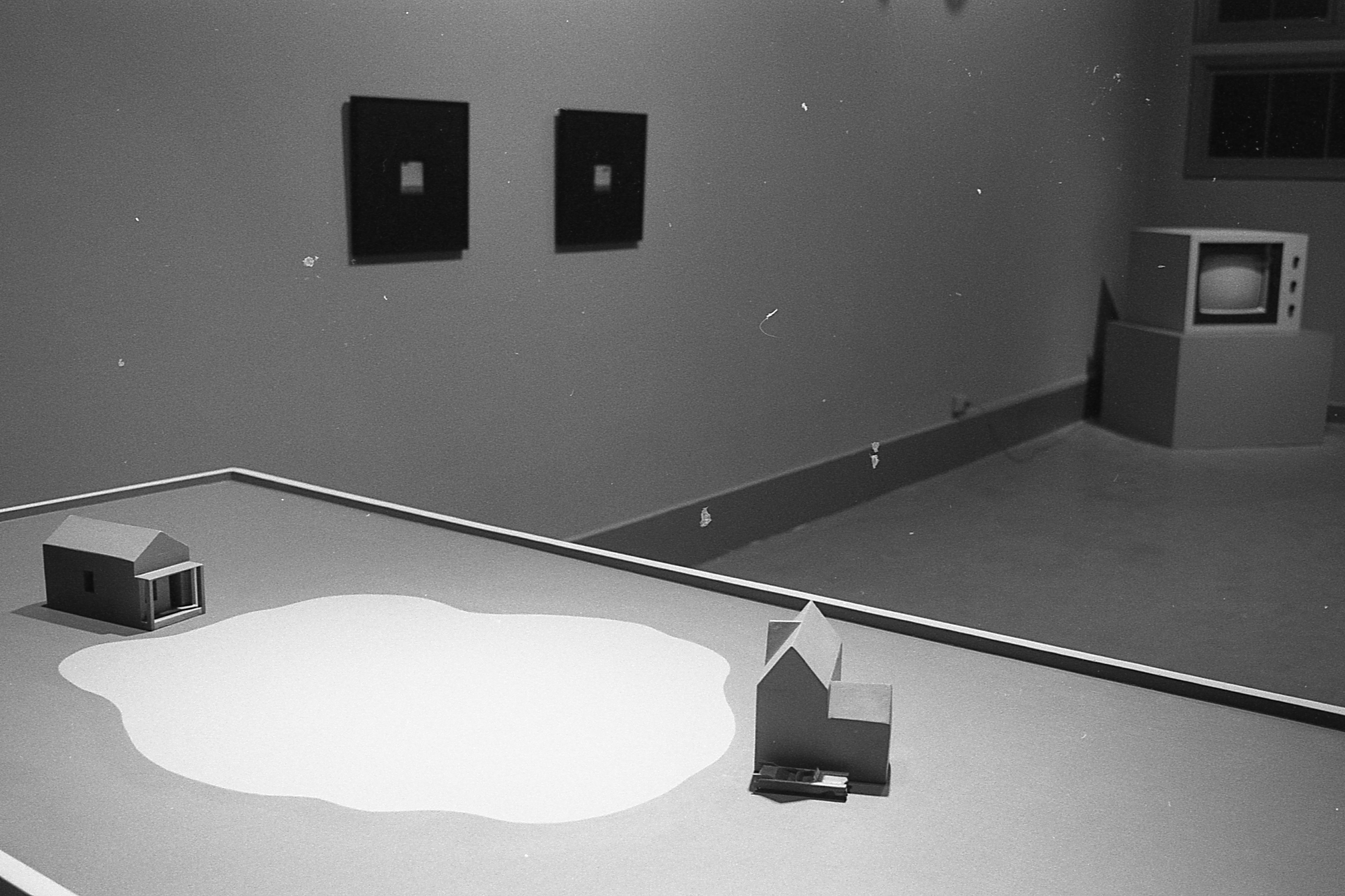 Small house-like wooden sculptures appear on a table with a large white image on the ground between them. A video monitor and framed images are visible in the background.