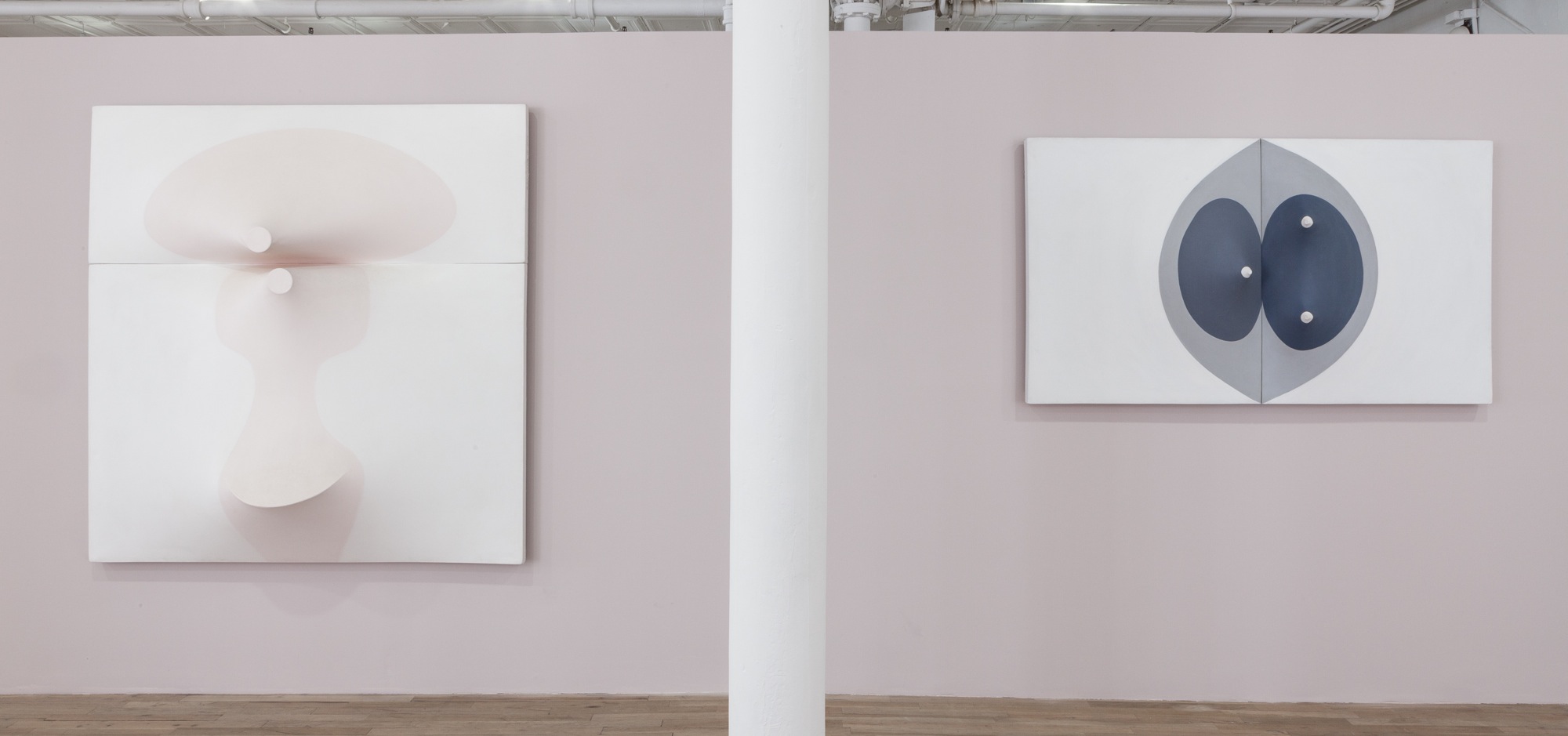 Two paneled works hang on either side of a white column on a light, mauve-colored wall. Both canvases are slightly three-dimensional, with elements that stretch in or out from their surfaces.