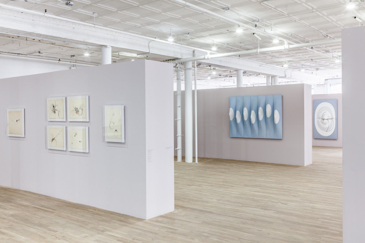 An angled view of three light, mauve-colored and parallel walls displaying drawings and paneled canvases in an exhibition space with wooden flooring. The canvases are slightly three-dimensional, with elements that stretch in or out from their surfaces.