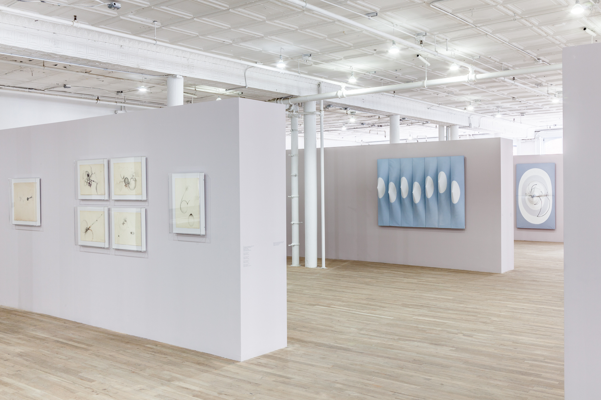 An angled view of three light, mauve-colored and parallel walls displaying drawings and paneled canvases in an exhibition space with wooden flooring. The canvases are slightly three-dimensional, with elements that stretch in or out from their surfaces.