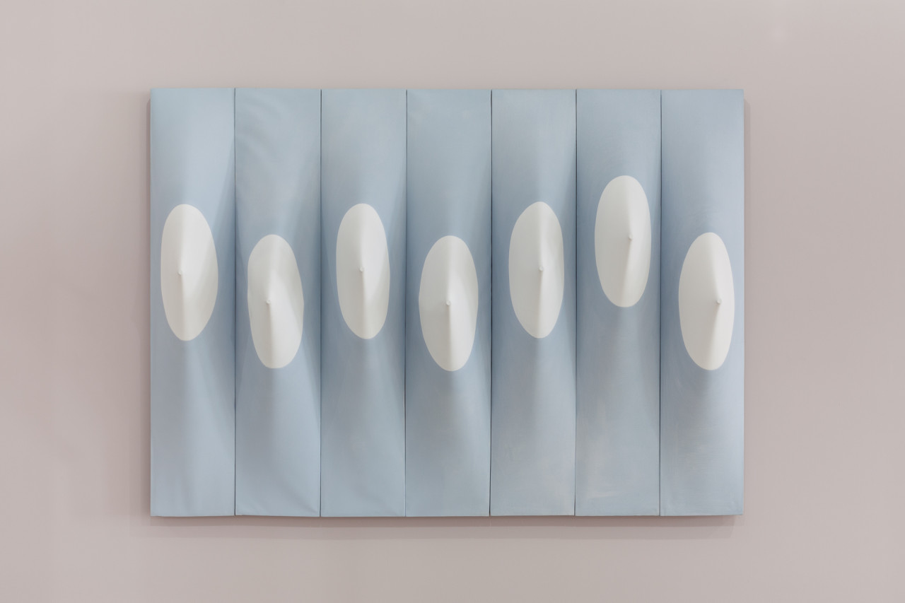 Seven vertical light blue panels, positioned side-by-side, displayed on a light mauve wall. Each panel contains a white circle positioned near its vertical center. Within each circle, the panel