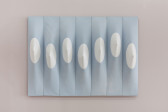 Seven vertical light blue panels, positioned side-by-side, displayed on a light mauve wall. Each panel contains a white circle positioned near its vertical center. Within each circle, the panel's canvas is stretched outward by a dowel.