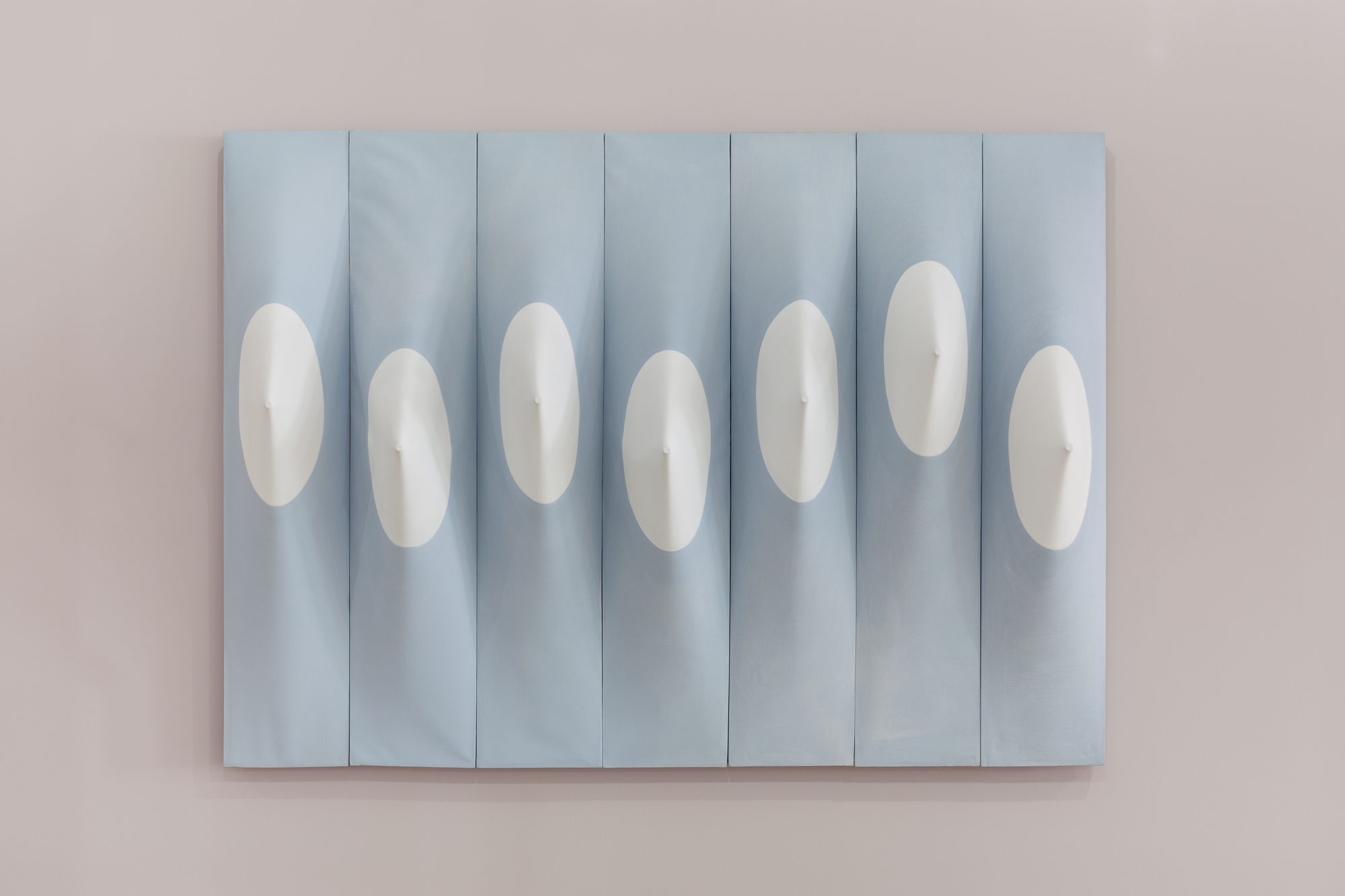 Seven vertical light blue panels, positioned side-by-side, displayed on a light mauve wall. Each panel contains a white circle positioned near its vertical center. Within each circle, the panel's canvas is stretched outward by a dowel.