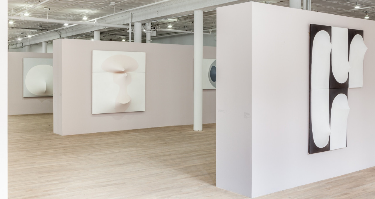 Three light, mauve-colored walls in an exhibition space each display several canvases. The three walls are arranged such that they are parallel to each other. Each of the canvases are slightly three-dimensional, with elements that stretch in or out from their surfaces.