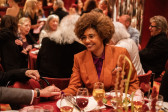 LaToya Ruby Frazier smiles while seated at a dinner table, holding hands with someone whose body is out of frame. She wears an orange suit jacket, a purple collared shirt, and a pocket square bearing a print of various orange flowers.