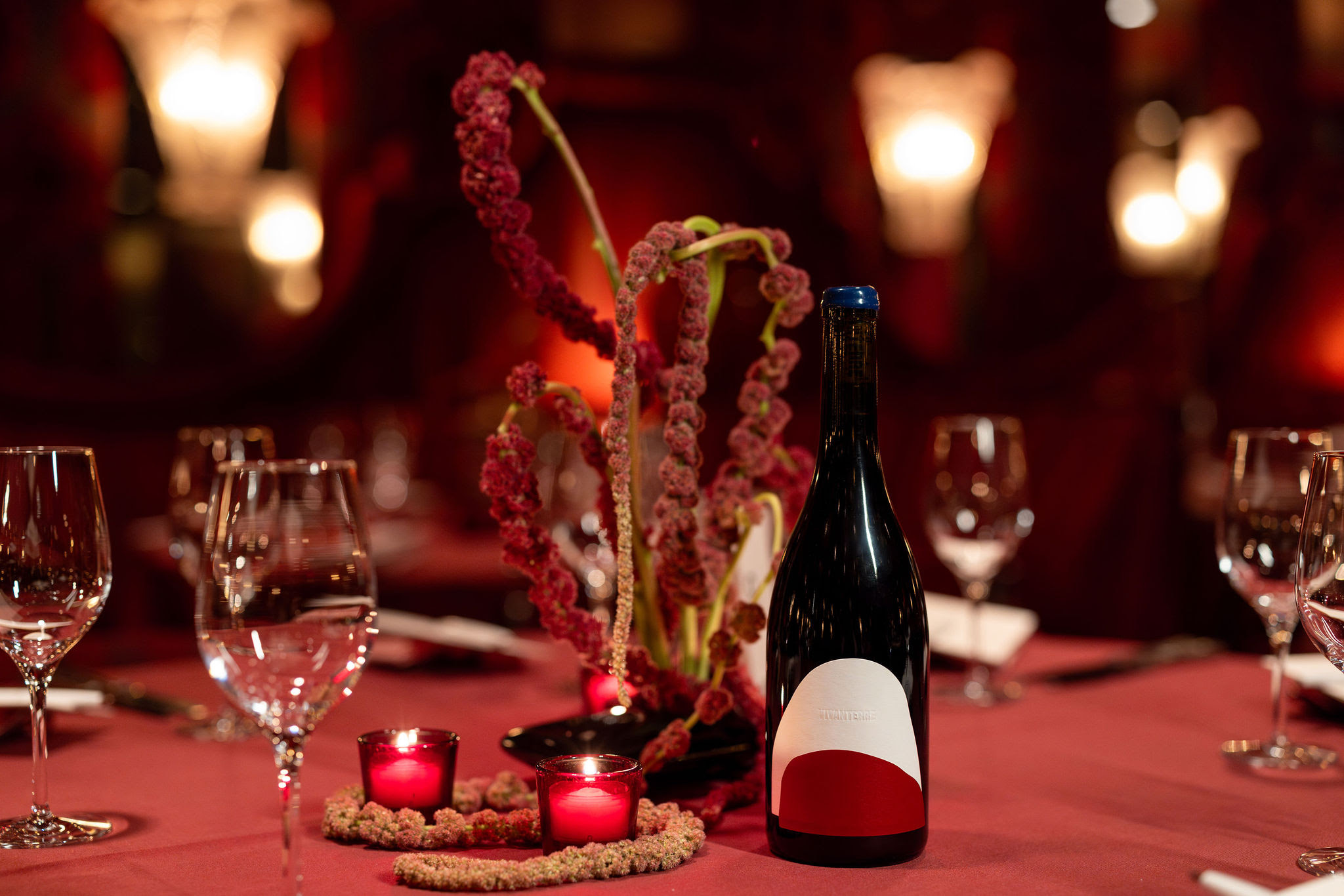 A bottle of wine sits on a set dining table covered in a red tablecloth, surrounded by wine glasses. The wine label features an irregular curved white semi-oval, overlaid with a smaller burgundy red shape, and the embossed text 