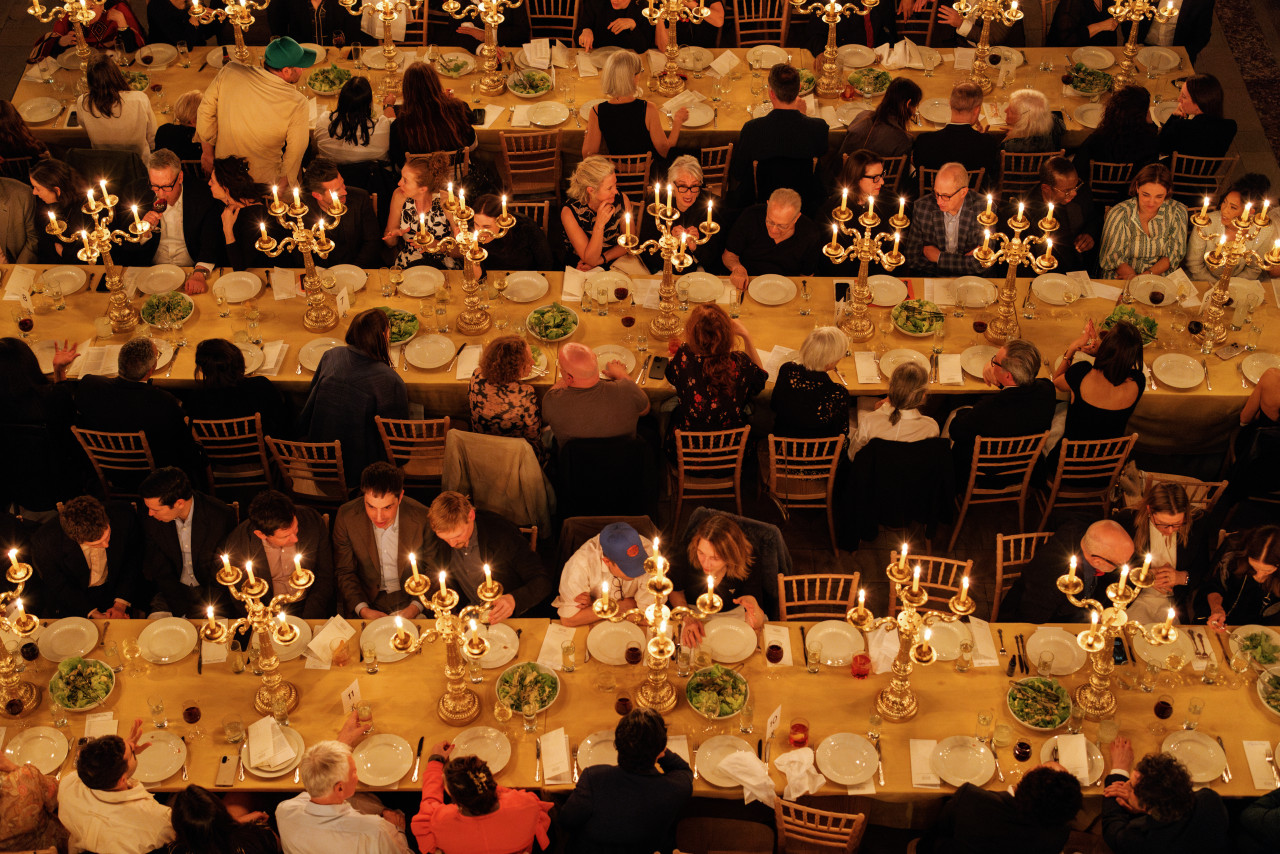 Aerial view of three long dining tables with rows of candlebras running down the middle of each. Rows of seated guests on each side of the tables are chatting and having dinner.