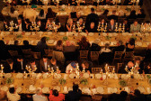 Aerial view of three long dining tables with rows of candlebras running down the middle of each. Rows of seated guests on each side of the tables are chatting and having dinner. 