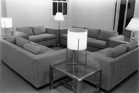 Black and white photo of four couches arranged in a square. Four lamps rest on glass side tables positioned at each corner of the square.