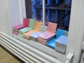 A series of booklets with monochrome covers sit in stacks on the interior of a windowsill. Each of the booklets' covers displays thin text spelling out various names, including 