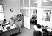 A black and white photo of an office with several people standing and sitting behind desks.