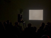 A film projector is set up in the middle of a darkened room, facing a blank wall. A rectangular grey image is projected onto the wall. A group of seated spectators are backlit by the projected image.