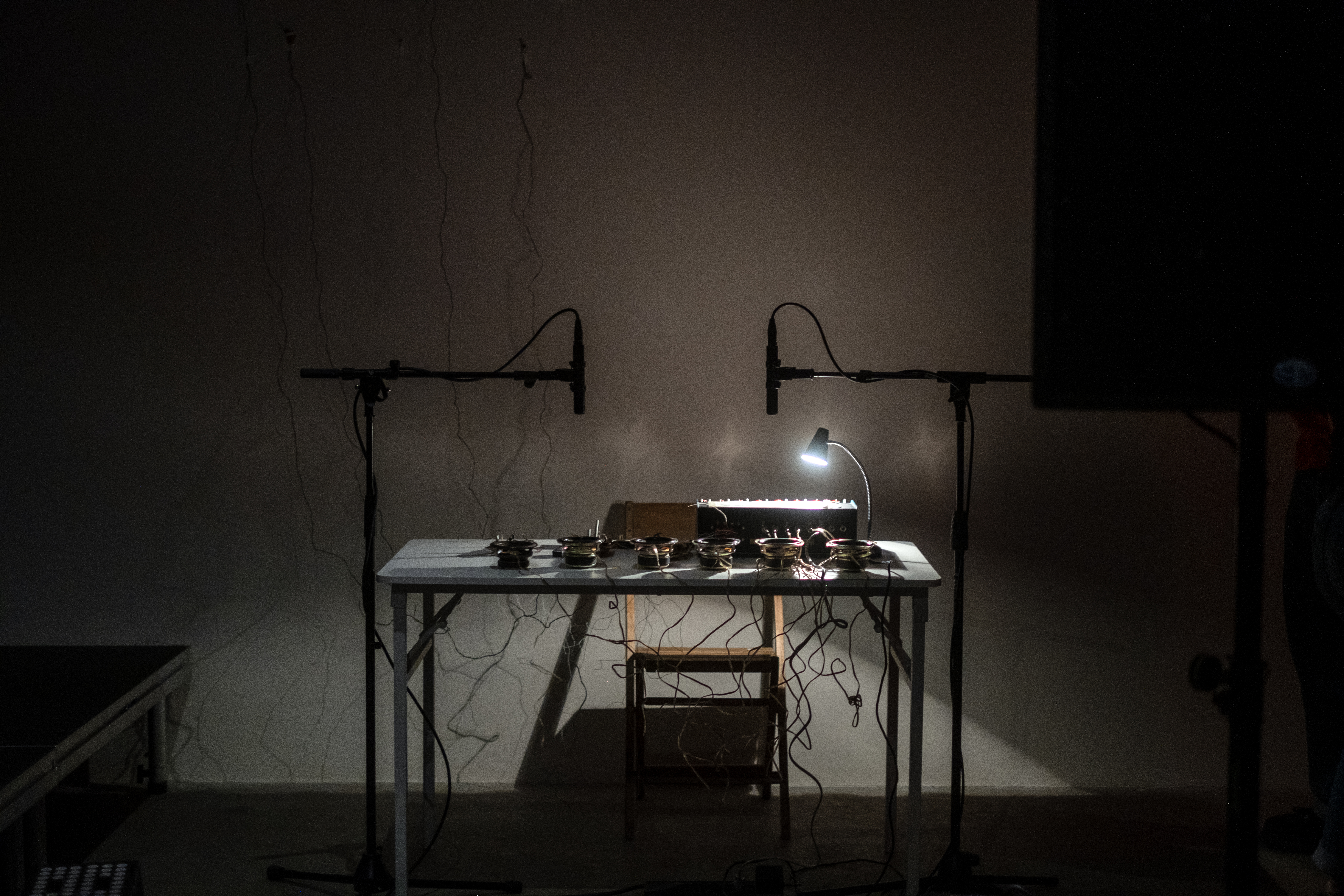 A color image shows a rectangular white desk in a darkened space. An audio device sits on the desk, surrounded by speakers and loose wires. A lamp illuminates the device’s controls, while two microphones hang above it, pointed downward. Behind the desk, wires from vertically mounted light sensors are visible in the shadows.