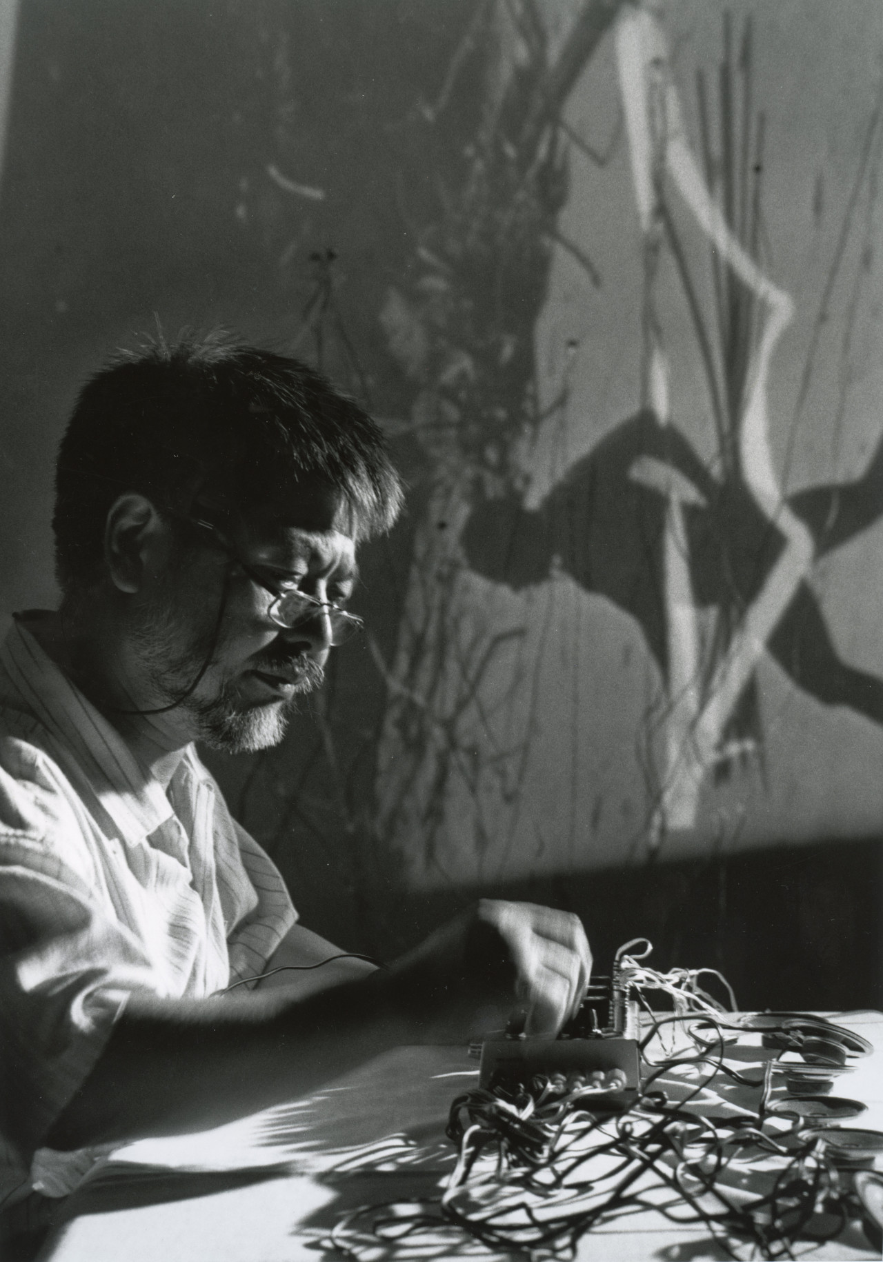 A black-and-white image shows a person seated behind a white desk, adjusting an audio device with their hands. The device is surrounded by replacement speakers with organically shaped wires. In the background, a projected video shows a landscape and a shadowy figure.