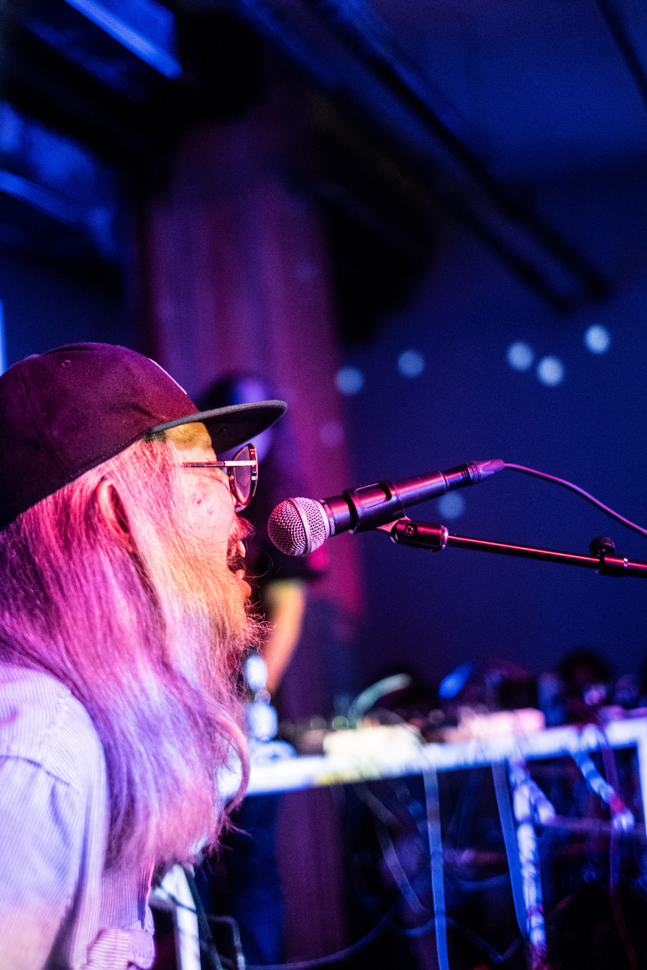 A figure leans into a microphone from the side with blue, purple, and pink projections cast onto them.