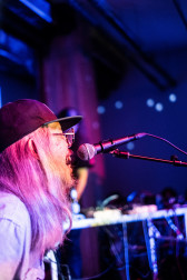 A figure leans into a microphone from the side with blue, purple, and pink projections cast onto them.