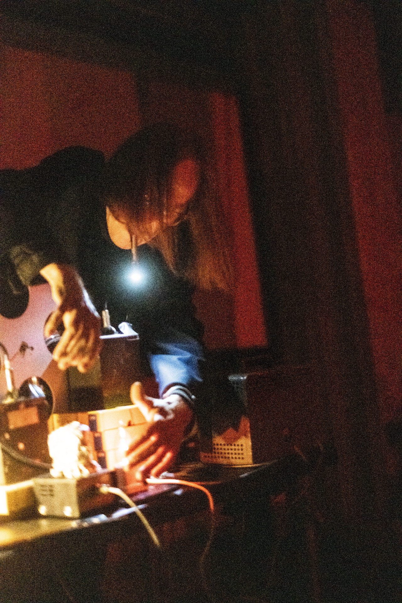 A figure leans over to arranges audio equipment on a desk using a flashlight held in their mouth. Red light fills the background.