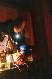 A figure leans over to arranges audio equipment on a desk using a flashlight held in their mouth. Red light fills the background.