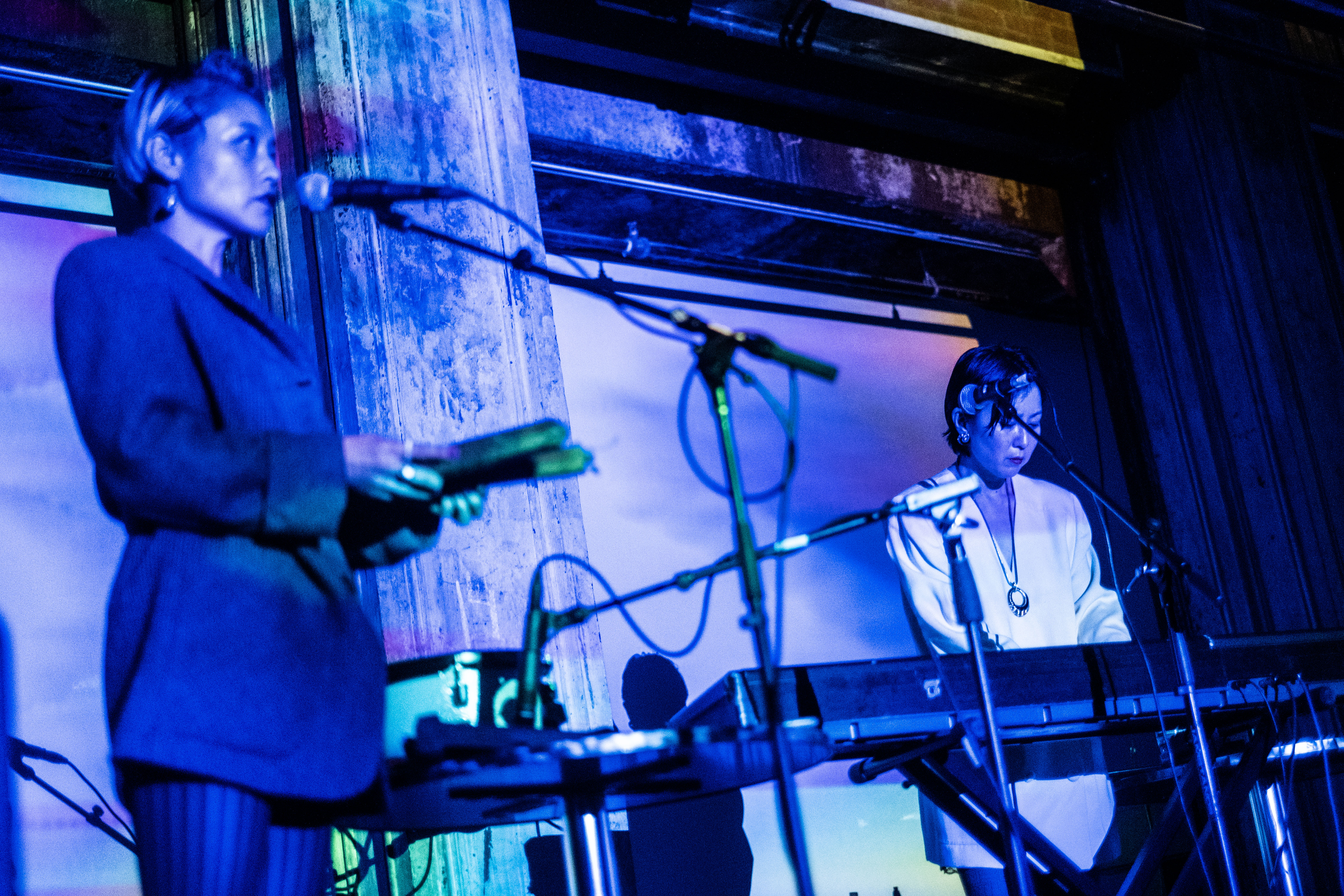 Two figures play music on a stage, the left figure plays a keyboard. Washes of blue and green light are projected onto them.