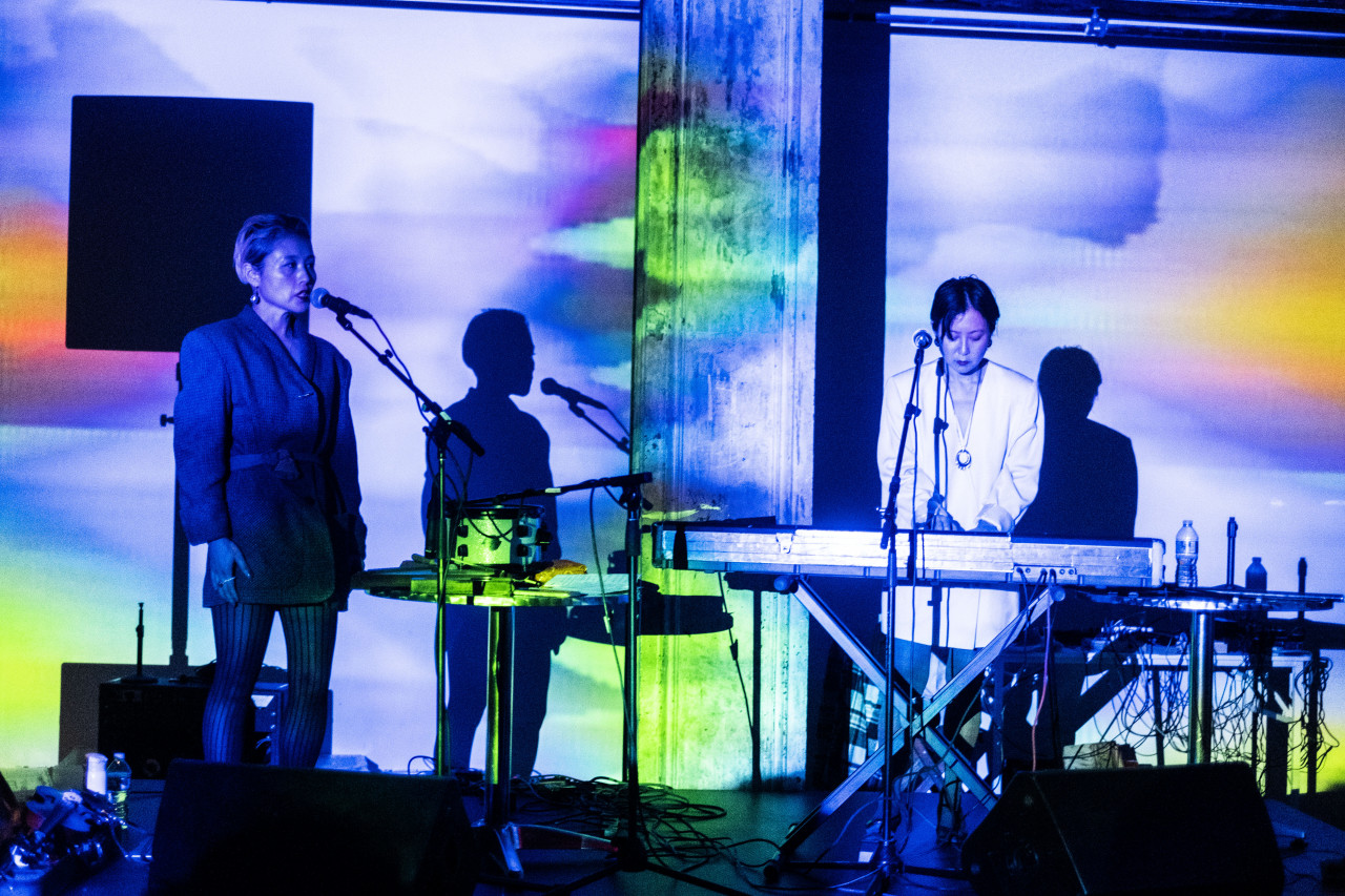 Two figures perform on a stage with a keyboard. Washes of blue, green, orange and pink light are projected onto them.