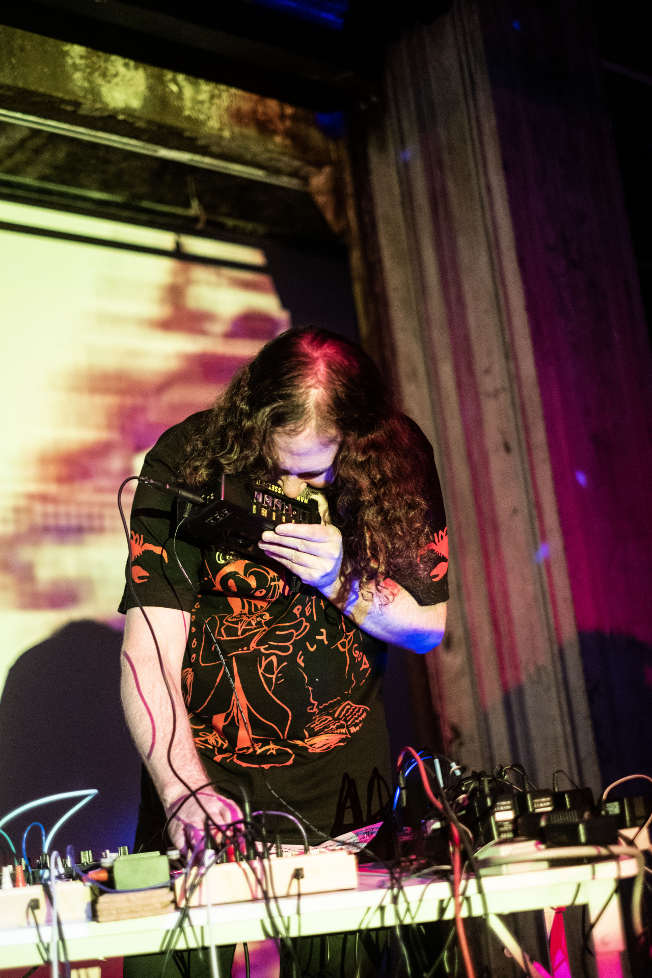 A figure performs on stage behind a table with audio equipment and holds an audio box to their mouth.