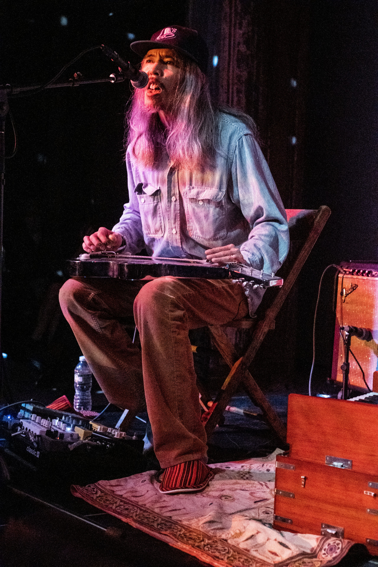 A figure performs on stage into a microphone with a small electric guitar in their lap.