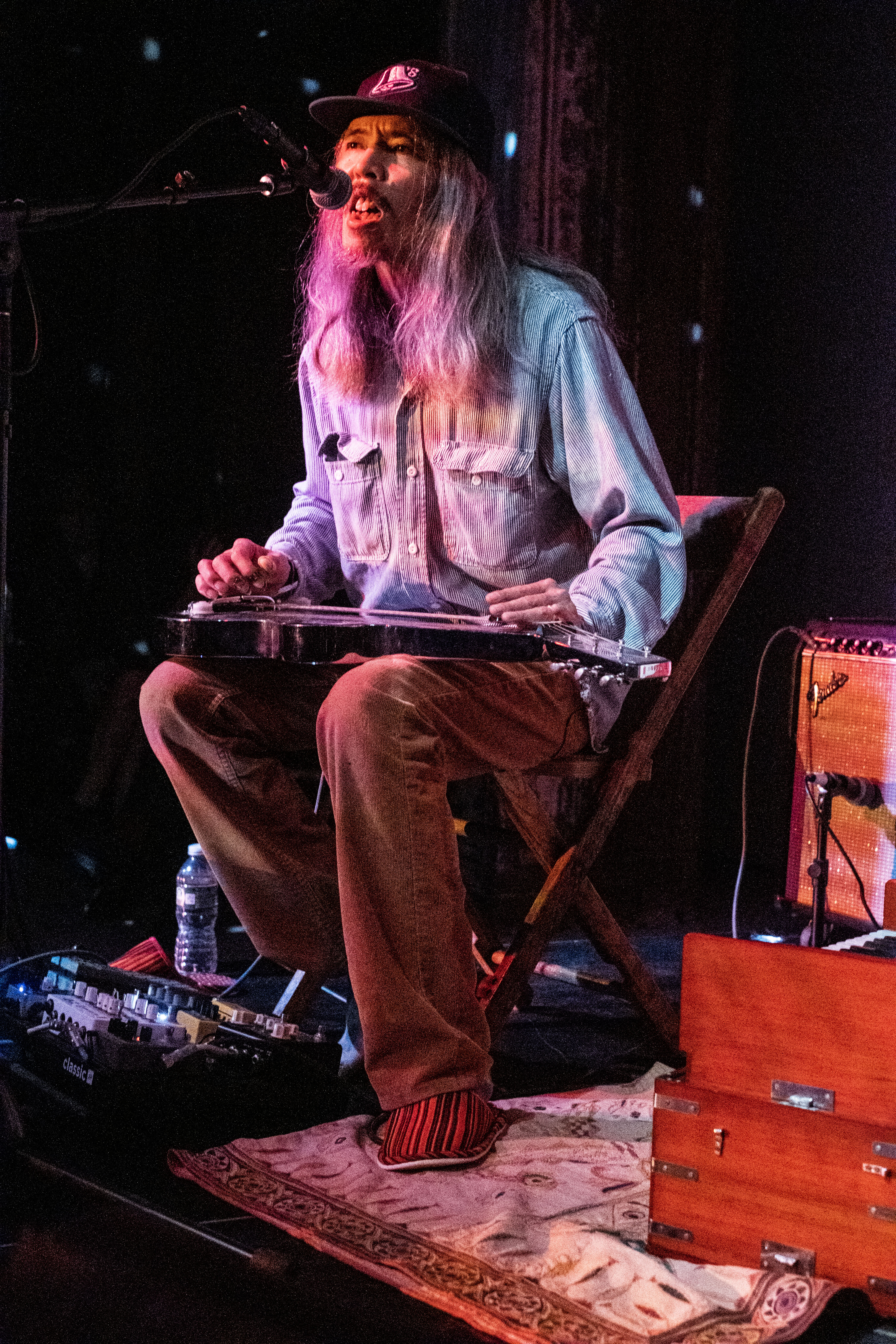 A figure performs on stage into a microphone with a small electric guitar in their lap.