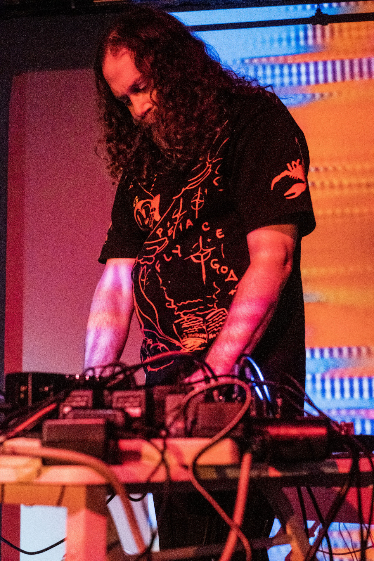 A figure stands in front of an orange and blue projection and performs with audio equipment.