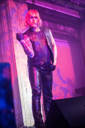 A figure dressed in leather performs on stage with a purple projection on them.