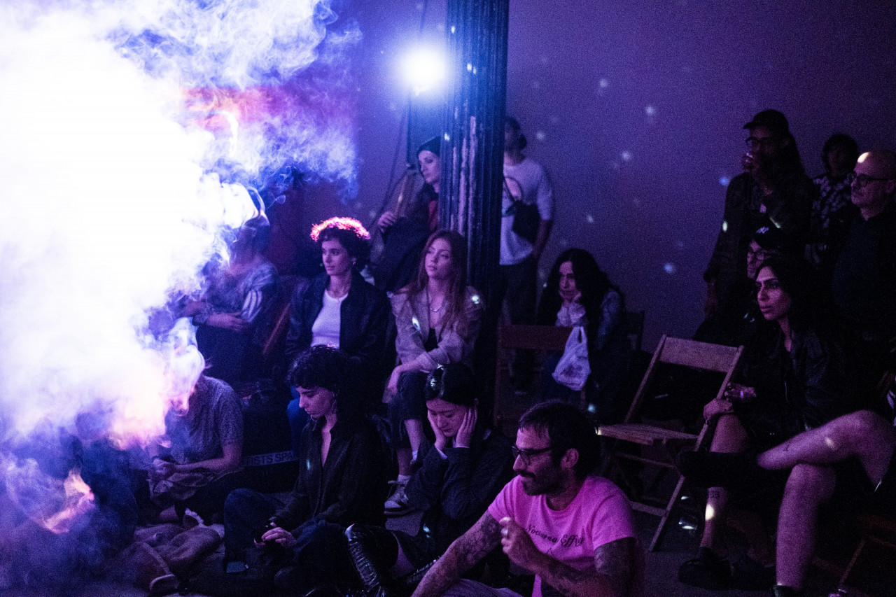 An audience watches a performance, smoke fills the room to the left and a projector casts dark blue light over the audience.
