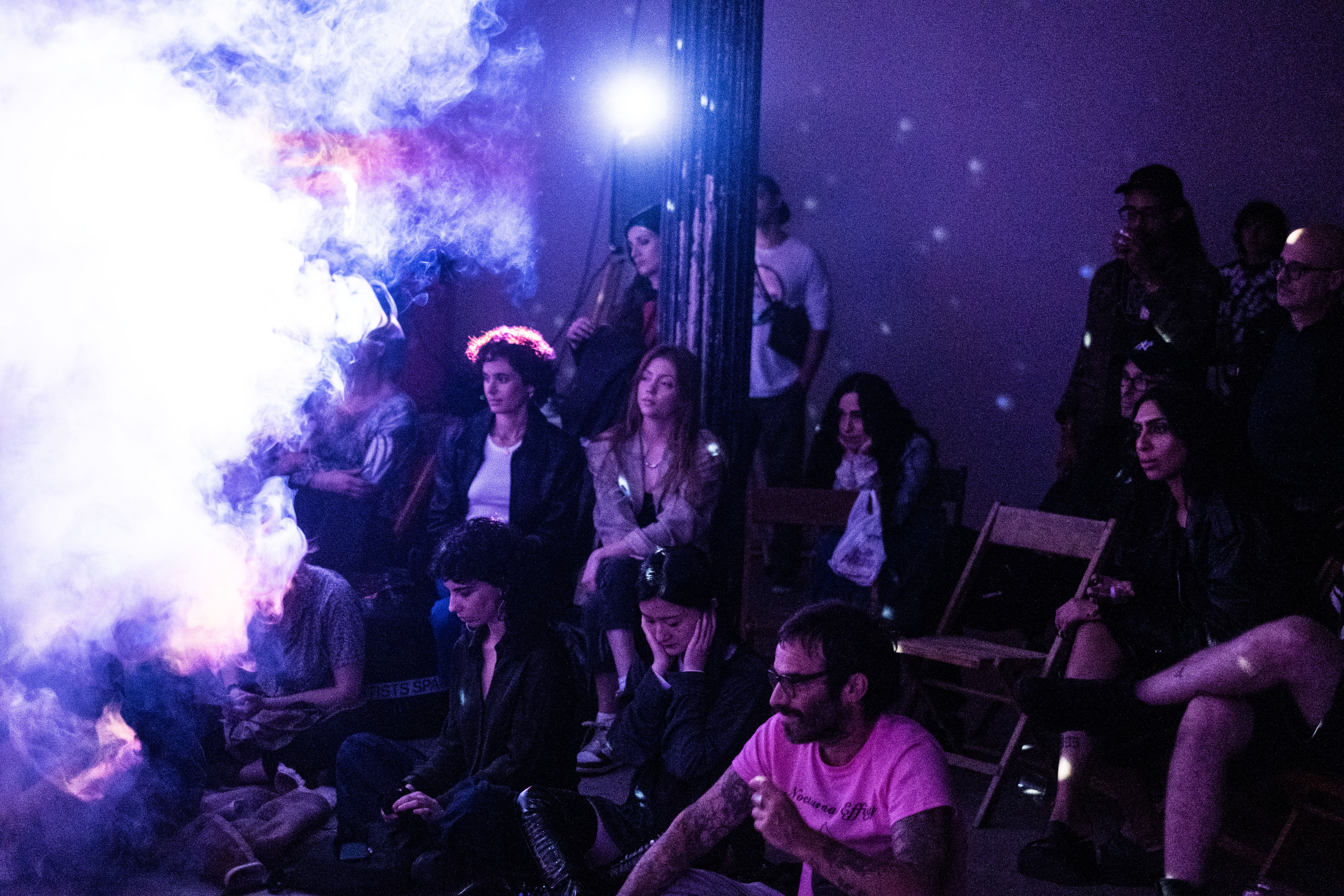 An audience watches a performance, smoke fills the room to the left and a projector casts dark blue light over the audience.