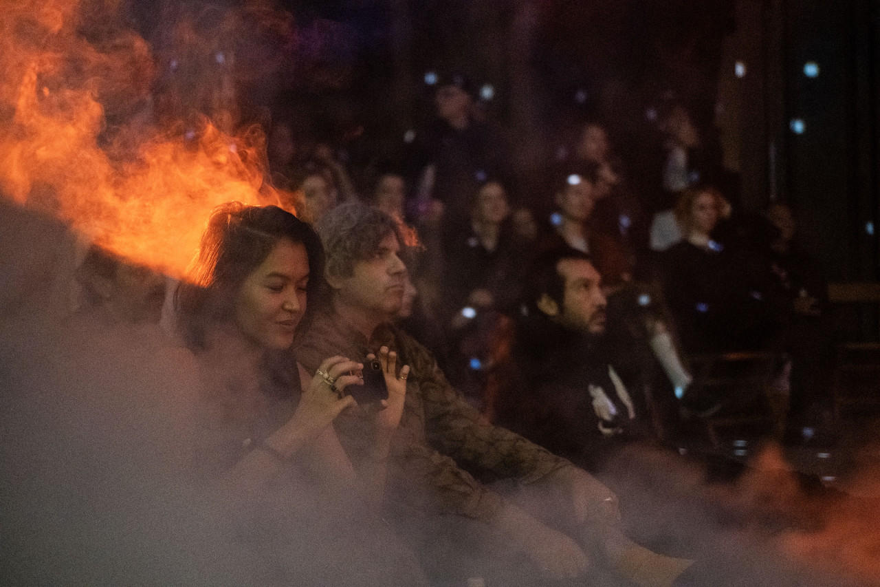 An audience watches a performance, smoke and an orange light fill the room.