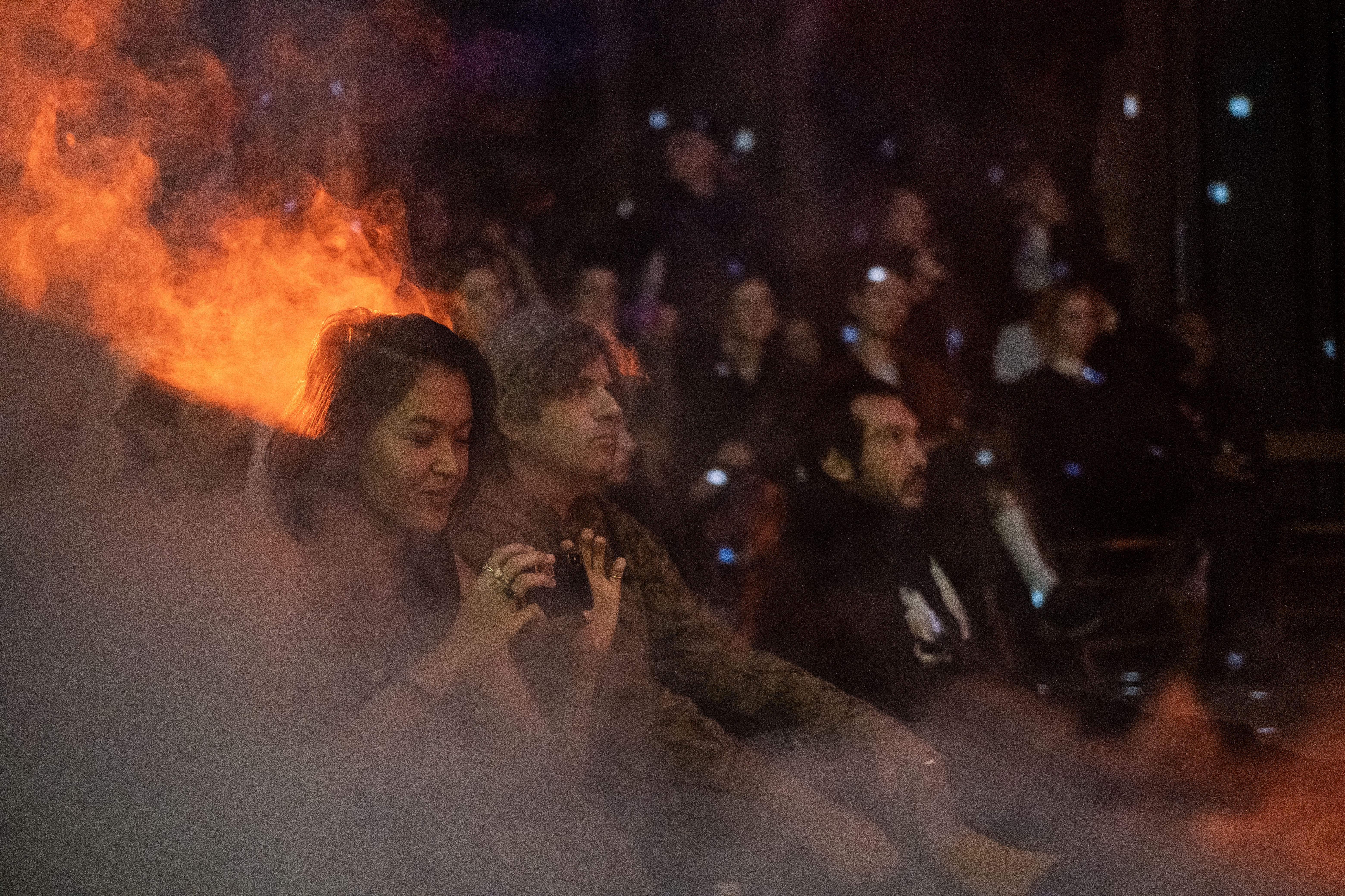An audience watches a performance, smoke and an orange light fill the room.