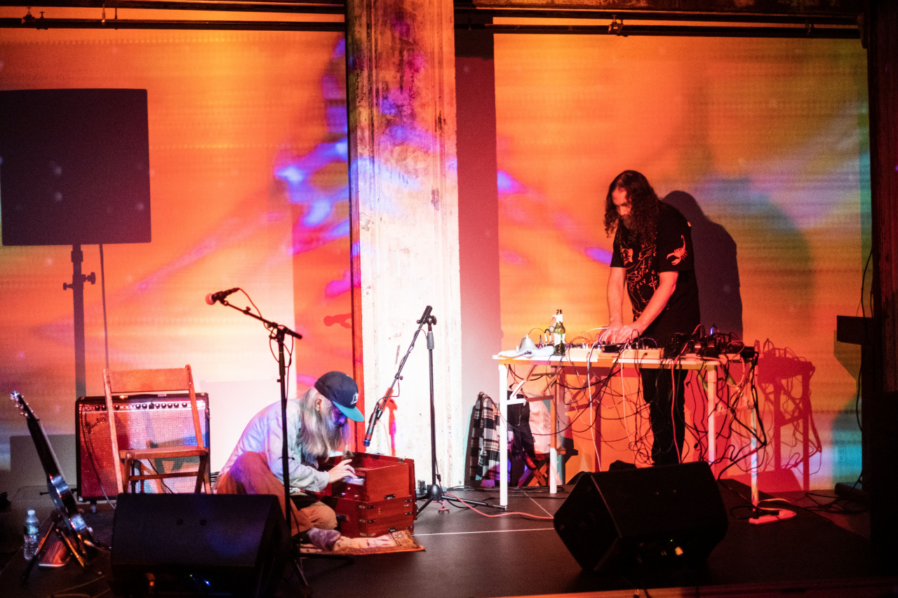 Two figures perform on stage. To the left a figure sits on the floor playing on a small wooden organ and to the right a figure stands behind a desk using a mixer. Orange and blue light are projected onto the stage.