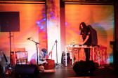 Two figures perform on stage. To the left a figure sits on the floor playing on a small wooden organ and to the right a figure stands behind a desk using a mixer. Orange and blue light are projected onto the stage.