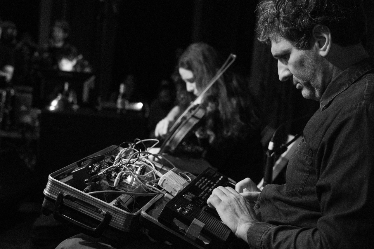 Black-and-white image of two figures performing on stage. On the right a figure looks down and manipulates audio equipment held in a briefcase. To the left and behind another figure is out of focus and plays the violin.