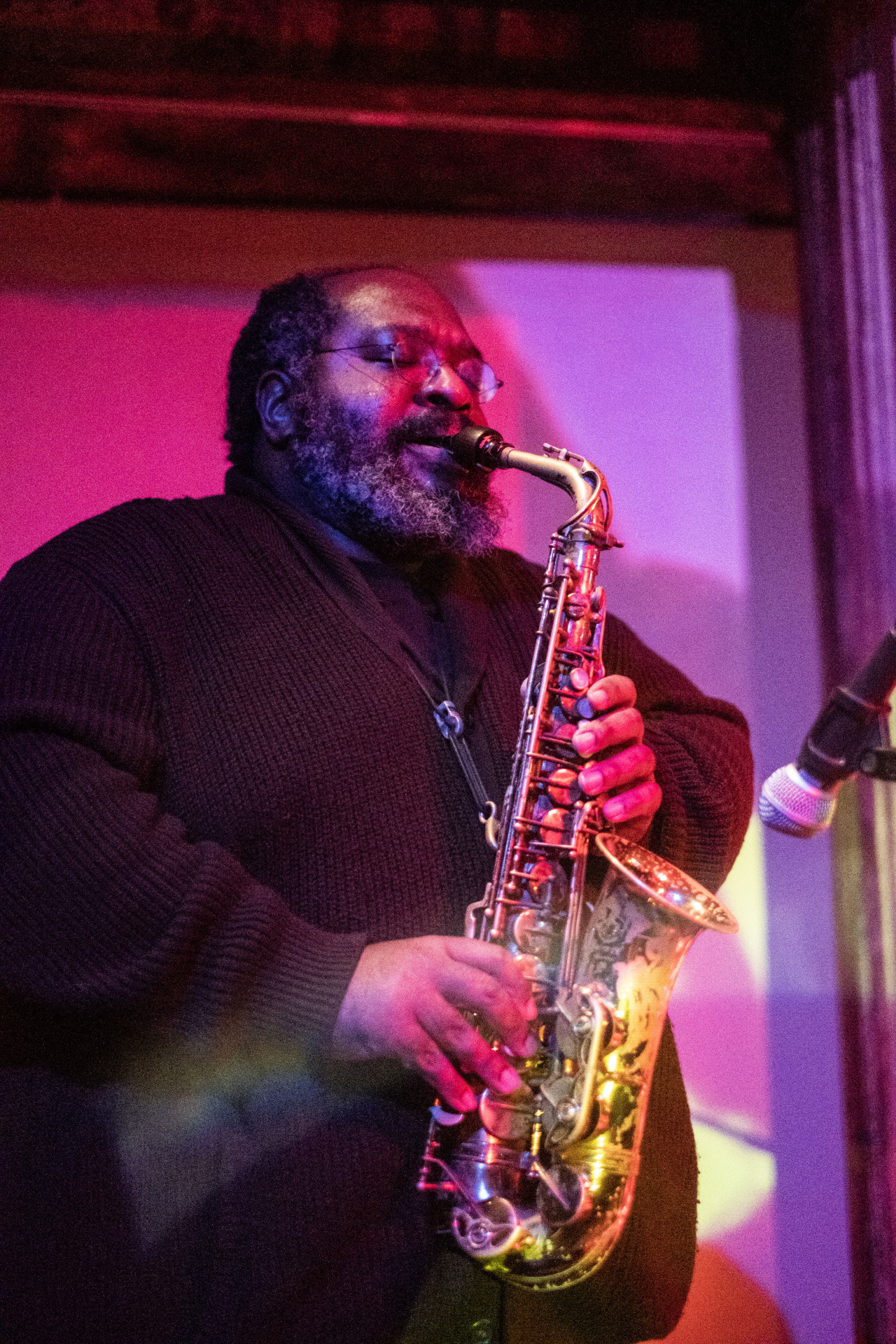 A figure plays the saxophone into a microphone. Red and purple light are cast onto them.