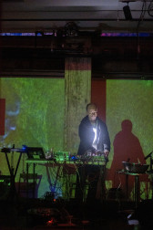 A figure in a black hoodie and an LED light stapped on their chest performs on a stage, behind a table full of audio equipment. A green light projection is cast onto the stage.