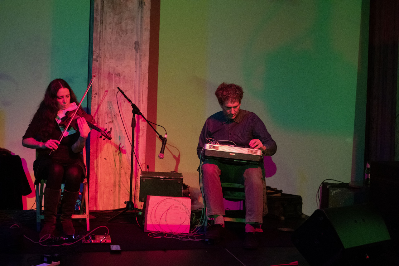 Two figures are seated on stage. The figure on the left plays the violin and the figure on the right holds a briefcase full of audio equipment.