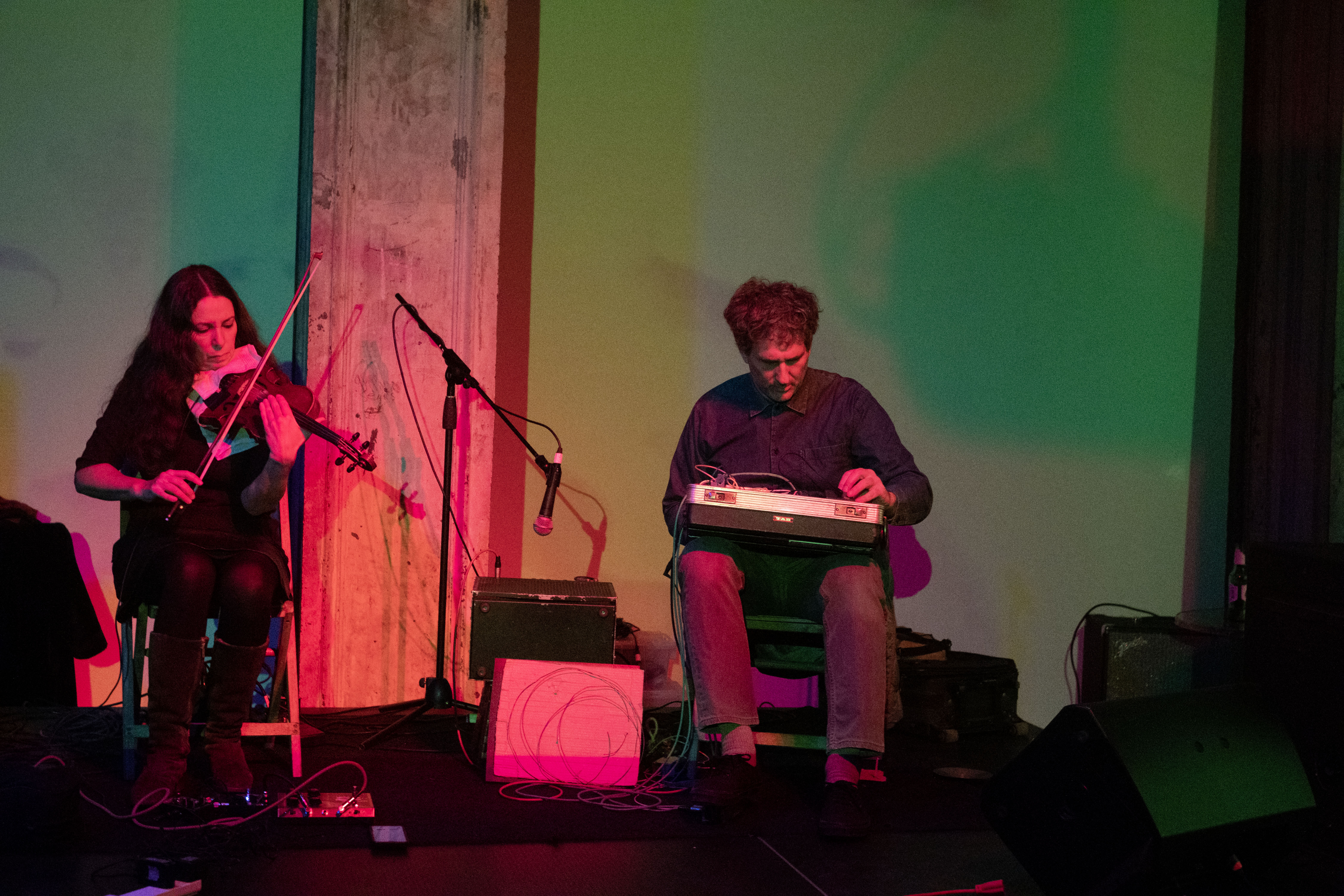 Two figures are seated on stage. The figure on the left plays the violin and the figure on the right holds a briefcase full of audio equipment.